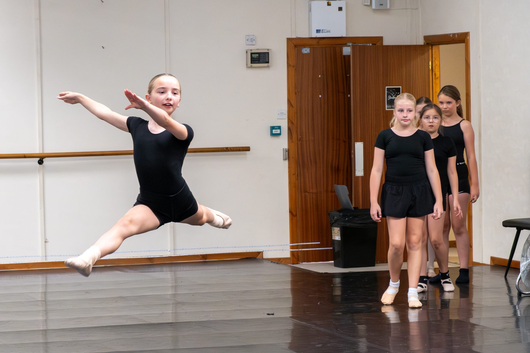 A Studio 19 dancer rehearsing with arms raised, fellow dancers mirroring her in the studio